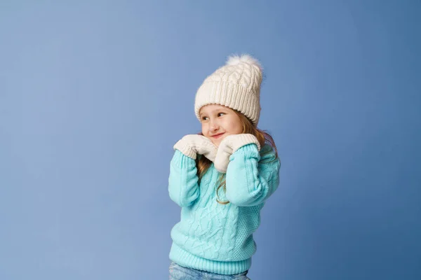 身穿白色针织帽子 毛衣和手套的可爱小女孩的双手紧握着脸 在蓝色的背景下微笑着 穿着保暖衣服的孩子把目光移开 复制空间 — 图库照片
