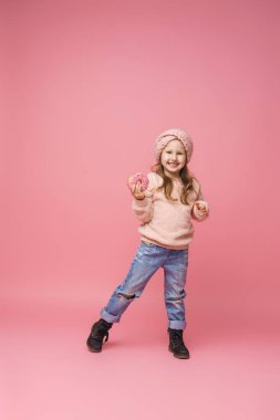 Tüylü kazak ve saç bandı takmış elinde donutla gülümseyen küçük bir kız. Tatlı aperatif, lezzetli hamur işleri. Bir çocuk, uzun bir stüdyoda pembe arka planda lezzetli bir tatlı gösterir.