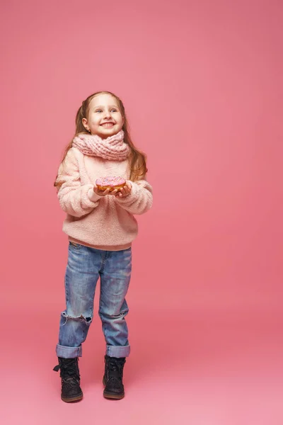 ふわふわのセーターとヘッドバンドの女の子がドーナツを手に笑っていました 甘い前菜 おいしいペストリー 子供はピンクの背景においしいデザートを見せてくれます — ストック写真