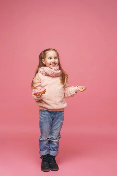 穿着毛绒绒毛衣 头戴头巾的小女孩 手里拿着甜甜圈 微笑着 甜开胃菜 好吃的糕点 一个孩子在一个粉红的背景上 在工作室里展示了一份美味的甜点 — 图库照片
