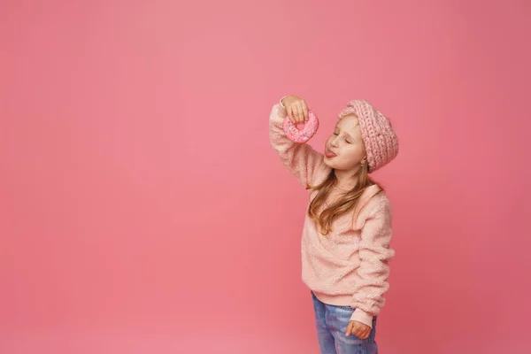 ふわふわのセーターとヘッドバンドの女の子がドーナツを手に笑っていました 甘い前菜 おいしいペストリー 子供はスタジオでピンクの背景においしいデザートを見せます — ストック写真