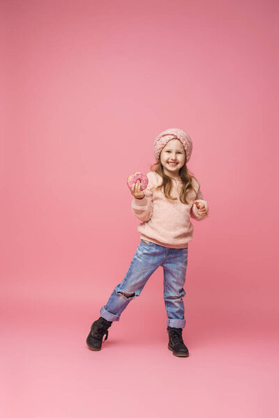 Маленькая девочка в пушистом свитере и повязке на голове, улыбающаяся с пончиком в руке. Сладкая закуска, вкусная выпечка. ребенок показывает вкусный десерт на розовом фоне в студии