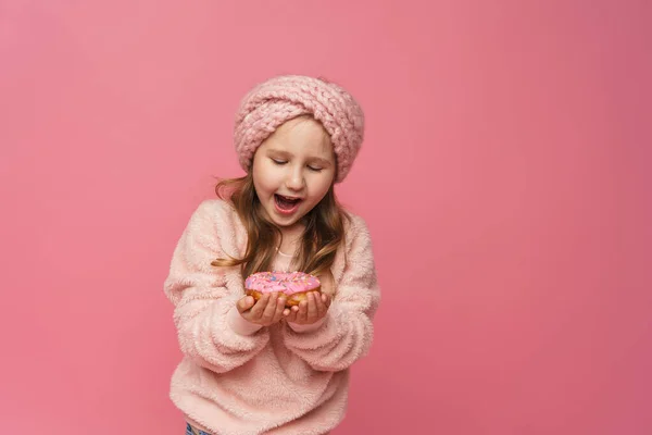 ふわふわのセーターとヘッドバンドの女の子がドーナツを手に笑っていました 甘い前菜 おいしいペストリー 子供はスタジオでピンクの背景においしいデザートを見せます — ストック写真