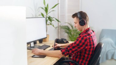 Yan görüş, kulaklıklı, kalem tableti kullanan genç bir adam evde bilgisayarın önünde rötuş yapıyor. Fotoğrafçı fotoğrafları inceler. Boş beyaz bilgisayar ekranı. Serbest çalışan biri. Uzaktan kumanda. Günlük tutma.