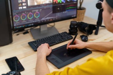 Genç kameraman, evdeki bilgisayardaki videoyu işlemek için kalem tableti ve özel program kullanıyor. Video düzenleme. Bloglar. Serbest çalışan biri. Uzaktan kumanda. Antrenman yapıyorum. Omzunun üstünden bak.