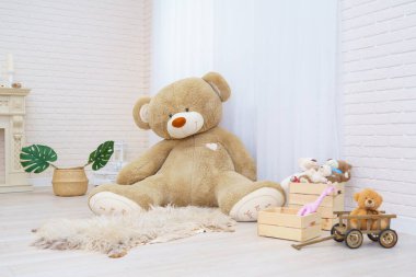 Dev oyuncak ayı, oyuncak sepetinde oyuncak, oyuncak ayılar, ahşap araba ve canavar yapraklarıyla dolu parlak bir odada oturuyor. Hafif tuğlalı duvarları ve ahşap parke döşemeleri olan modern bir iç mekan. Boşluğu kopyala