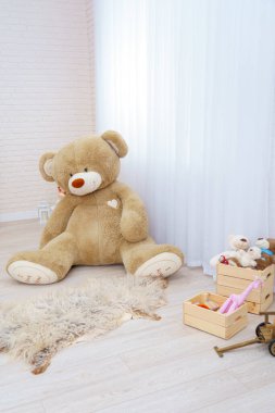Dev oyuncak ayı, oyuncak sepetinde oyuncak, oyuncak ayılar, ahşap araba ve canavar yapraklarıyla dolu parlak bir odada oturuyor. Hafif tuğlalı duvarları ve ahşap parke döşemeleri olan modern bir iç mekan. Boşluğu kopyala
