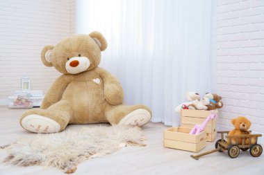 Dev oyuncak ayı, oyuncak sepetinde oyuncak, oyuncak ayılar, ahşap araba ve canavar yapraklarıyla dolu parlak bir odada oturuyor. Hafif tuğlalı duvarları ve ahşap parke döşemeleri olan modern bir iç mekan. Boşluğu kopyala