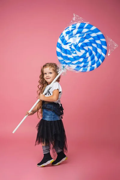 时尚的小女孩6岁 卷曲的头发 穿着时髦衣服的蓝白相间的大棒棒糖 全身上下都是粉色背景 小孩笑了一个情绪化的形象复制空间 — 图库照片