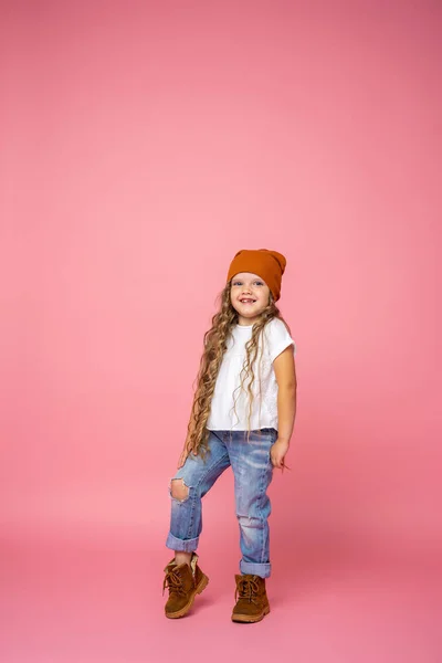 时尚的6岁女孩 穿着撕破的牛仔裤 头戴卷发的帽子 全身而退 与粉色背景隔离 动态的情感形象 复制空间 — 图库照片