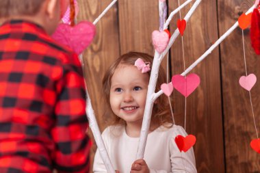 Sevgililer Günü 'nde çekilecek fotoğraf çekimi için arka plandaki mutlu küçük kız kırmızı ve pembe kalpli bir ağacın arkasından dışarı bakar. Neşeli bir çocuk Sevgililer Günü 'nü kutluyor