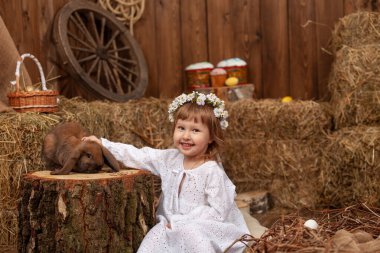 Easter decoration. cute little girl petting and feeding lop-eared rabbit. child in white dress and wreath flowers on his head, is in contact, communicates with pet rabbit, in hay barn on Easter day.