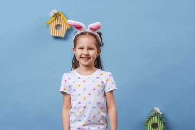 Şirin, gülümseyen küçük bir kız stüdyoda mavi bir arka planda duruyor ve elinde paskalya yumurtaları tutuyor. Komik, mutlu bebek, Paskalya günü tavşan kulağı takıyor. Paskalya yumurtası avında iyi eğlenceler. Boşluğu kopyala.