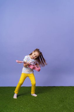 4 yaşında, şık, parlak elbiseli neşeli, beyaz bir kız pembe bir gitar çalıyor. Çocuk bir müzik aleti çalıyor ve eflatun arka planda ve yeşil çimlerde şarkı söylüyor..