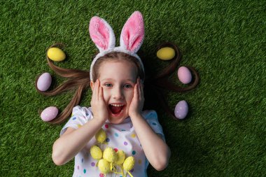 Duygusal küçük kız boyalı yumurtalarla çimenlerde uzanıyor. Saçında paskalya yumurtası var. Tavşan kulaklı çocuk ellerini yüzüne dayayıp Paskalya günü bağırıyor. Neşeli küçük kız Paskalya yumurtalarını tutuyor.