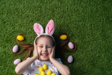 Duygusal küçük kız boyalı yumurtalarla çimenlerde uzanıyor. Saçında paskalya yumurtası var. Tavşan kulaklı çocuk ellerini yüzüne dayayıp Paskalya günü bağırıyor. Neşeli küçük kız Paskalya yumurtalarını tutuyor.