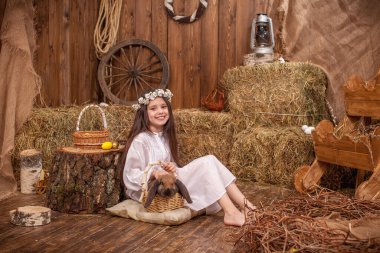 Paskalya süsü. Tatlı kız sarkık kulaklı tavşanı okşayıp okşuyor. Beyaz elbiseli çocuk ve başında çiçek çelengi, temas halinde, evcil tavşanla iletişim kuruyor, saman ahırında oturuyor.
