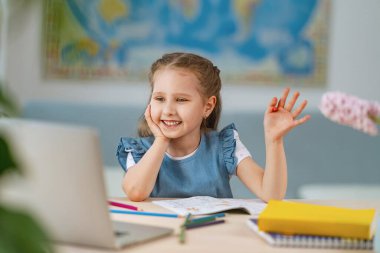 Çevrimiçi çalışmak için dizüstü bilgisayar kullanan zeki küçük bir kız, uzaktan eğitim öğretmenini selamlıyor, bir soruyu cevaplıyor. etkileşimli E-Eğitim Mesafesi Ev Öğrenimi.
