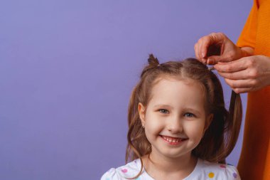 Anne, kızının uzun saçlarını mor arka planda örüyor. Beş yaşında bir kız oynuyor ve annesi saçını örüyor..
