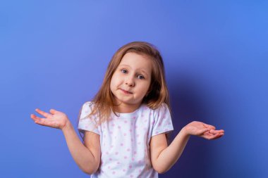 Küçük, üzgün, sarışın bir çocuk. 5 yaşında, tişörtlü suçlu bir kız. Stüdyonun mor arka planına el sallıyor. Çocuk yaşam tarzı kavramı. Kötü davranış..