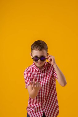 suspicious boy in dark sunglasses and a red checked shirt looks doubtfully at the camera., standing against the yellow background of the studio. Beckons with a finger