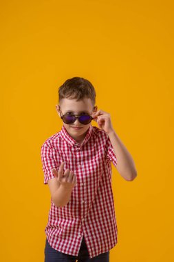 suspicious boy in dark sunglasses and a red checked shirt looks doubtfully at the camera., standing against the yellow background of the studio. Beckons with a finger