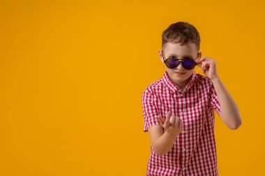 suspicious boy in dark sunglasses and a red checked shirt looks doubtfully at the camera., standing against the yellow background of the studio. Beckons with a finger