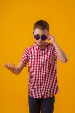 suspicious boy in dark sunglasses and a red checked shirt looks doubtfully at the camera., standing against the yellow background of the studio. Distrust of others