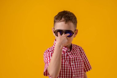 suspicious boy in dark sunglasses and a red checked shirt looks doubtfully at the camera., standing against the yellow background of the studio. Observes the environment