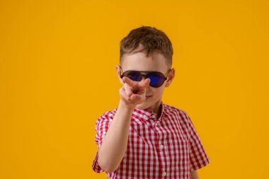 suspicious boy in dark sunglasses and a red checked shirt looks doubtfully at the camera., standing against the yellow background of the studio. Observes the environment