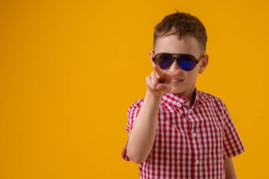 suspicious boy in dark sunglasses and a red checked shirt looks doubtfully at the camera., standing against the yellow background of the studio. Observes the environment