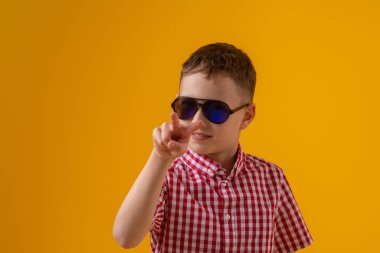 suspicious boy in dark sunglasses and a red checked shirt looks doubtfully at the camera., standing against the yellow background of the studio. Observes the environment