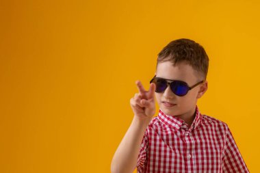 suspicious boy in dark sunglasses and a red checked shirt looks doubtfully at the camera., standing against the yellow background of the studio. Observes the environment