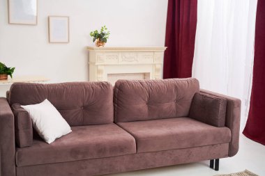 Büyük kahverengi bir kanepesi, modern bir hasır halısı, bir paspası ve büyük mumları olan bir şöminesi olan parlak, rahat bir ev odası. Büyük pencere tül ve perdeler ile çerçevelenmiş, karartma perdeleri.