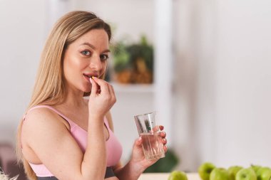 Hamile kadın elinde cam su ve vitaminlerle dizüstü bilgisayarla masada oturuyor. Kalsiyum ve folik asit alır, virüslere karşı korur. Dokunulmazlığı korumak için. Hipovitaminozu önleme