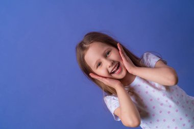 5 yaşında, tişörtünde siyah benekli sarı saçlar olan küçük bir kız gülümsüyor ve mor arka planda ellerini yüzüne dayıyor. Model eğleniyor ve şaşkına dönmüş hissediyor..