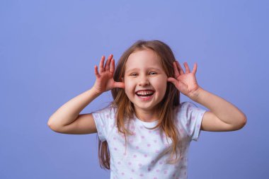 5 yaşında, sarı saçlı, tişörtlü, siyah benekli, mor arka planda mutlu bir şekilde gülümseyen küçük bir kız. Yakın plan. Model eğleniyor ve şaşkına dönmüş hissediyor..