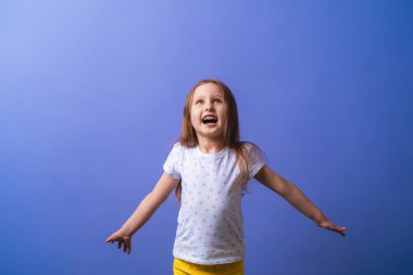 5 yaşında, sarı saçlı, siyah noktalı tişörtlü küçük bir kız kollarını yana açıyor ve mor bir arka planda mutlu bir şekilde gülümsüyor. Yakın plan.