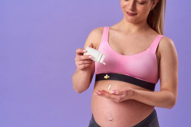 Hamile atletik kadın plastik kavanoz hapları tutuyor, vitamin ve mineraller alıyor. Mor arka planda dururken kapsülleri eline döküyor. Sağlıklı hamilelik, sağlık desteği. Yakın plan. Boşluğu kopyala