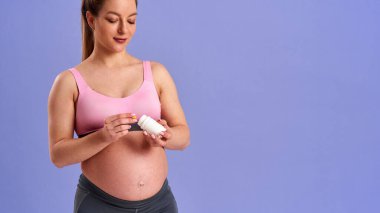 Hamile atletik kadın plastik kavanoz hapları tutuyor, vitamin ve mineraller alıyor. Mor arka planda dururken kapsülleri eline döküyor. Sağlıklı hamilelik, sağlık desteği. Yakın plan. Boşluğu kopyala