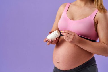 Hamile atletik kadın plastik kavanoz hapları tutuyor, vitamin ve mineraller alıyor. Mor arka planda dururken kapsülleri eline döküyor. Sağlıklı hamilelik, sağlık desteği. Yakın plan. Boşluğu kopyala