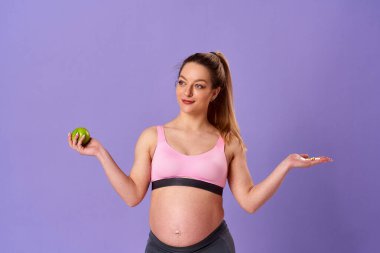 Hamile atletik kadın bir elinde haplar, diğer elinde yeşil elma, mor arka planda duruyor. Hamile sarışın hangi vitamini alacağına karar verir. Sağlıklı hamilelik, destek.