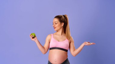 Hamile atletik kadın bir elinde haplar, diğer elinde yeşil elma, mor arka planda duruyor. Hamile sarışın hangi vitamini alacağına karar verir. Sağlıklı hamilelik, destek.
