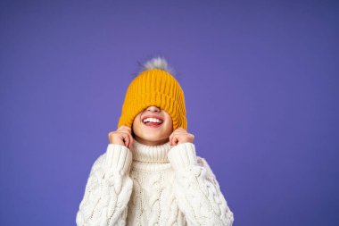 Örgü ören süveter giyen mutlu, neşeli bir çocuk mor arka planda sarı örülmüş bir şapkayı gözlerine çekiyor. Çocuk parlak kalın giysiler giydi ve kışın başlamasından mutlu. Boşluğu kopyala.