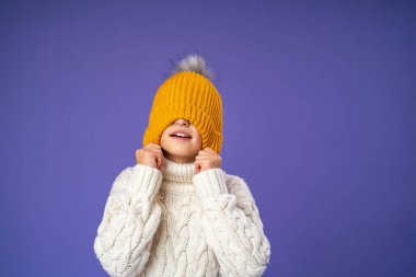 Örgü ören süveter giyen mutlu, neşeli bir çocuk mor arka planda sarı örülmüş bir şapkayı gözlerine çekiyor. Çocuk parlak kalın giysiler giydi ve kışın başlamasından mutlu. Boşluğu kopyala.