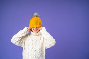 Örgü ören süveter giyen mutlu, neşeli bir çocuk mor arka planda sarı örülmüş bir şapkayı gözlerine çekiyor. Çocuk parlak kalın giysiler giydi ve kışın başlamasından mutlu. Boşluğu kopyala.