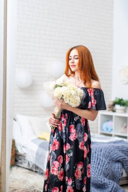 Güzel, genç bir kadın bir buket beyaz gülle parlak bir iç güzellikte çiçeklere bakıyor. Kıvırcık kızıl saçlı, uzun elbiseli, elinde çiçeklerle gülümseyen güzel bir kız. bahar festivali