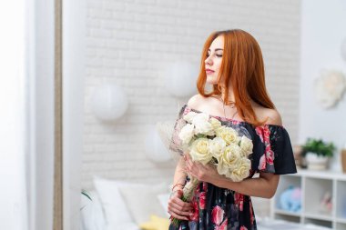 Güzel, genç bir kadın bir buket beyaz gülle parlak bir iç güzellikte çiçeklere bakıyor. Kıvırcık kızıl saçlı, uzun elbiseli, elinde çiçeklerle gülümseyen güzel bir kız. bahar festivali