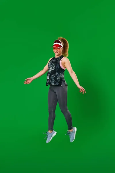 pregnant athletic woman in sportswear and a visor jumps on a green background. A pregnant blonde is engaged in sports. Healthy pregnancy, support. Sports during pregnancy. Close up, copy space.