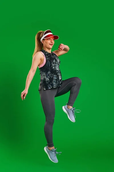 pregnant athletic woman in sportswear and a visor jumps on a green background. A pregnant blonde is engaged in sports. Healthy pregnancy, support. Sports during pregnancy. Close up, copy space.